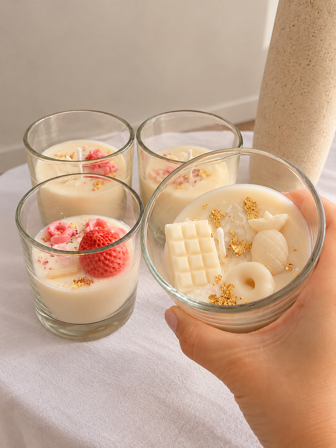 Mano sosteniendo una vela de cera con chocolate blanco, ananá y rosquilla, junto a otras velas con flores y frutilla